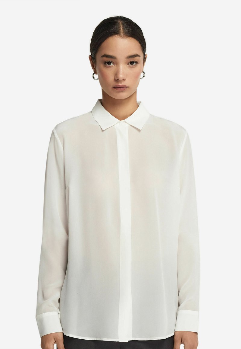 Jonge vrouw met donker haar in een lage knot, draagend zilveren oorbellen en een lange mouw witte blouse met kraag tegen een witte achtergrond.