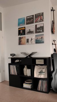 Black bookshelf with vinyl records, ceramic vases, a bag, and an amplifier. Wall displays various album covers; wooden floor visible.