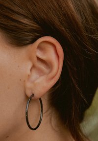 Schwarzer Metall-Hoop-Ohrstecker, zirkuläre Form, glatte Textur, hängt vom Ohr herab und betont die Konturen des Ohrs sowie das umliegende Haar.