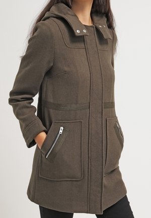 Femme portant un manteau à capuche marron mi-long avec des poches frontales zippées et une fermeture à boutons-pression au col. Mains dans les poches.