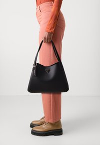 Borsa in pelle nera con design curvo, accessori in oro e un'etichetta con il logo. Indossata con pantaloni rosa chiaro e scarpe beige con lacci.