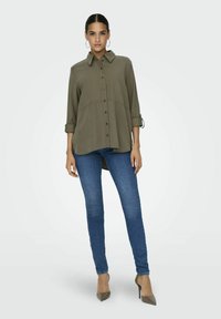 Blusa verde oliva con bottone frontale, colletto e maniche arrotolate, abbinata a jeans slim-fit blu e tacchi alti color taupe. Tessuto liscio.