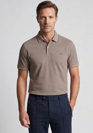 Man draagt een taupekleurig poloshirt met korte mouwen en donkerblauwe broek, staand tegen een effen lichte achtergrond.