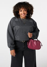 Vrouw met krullend haar draagt een donkergrijze trui met hartpatroon, zwarte broek, gouden sieraden en houdt een kleine bordeauxrode handtas vast voor een witte achtergrond.