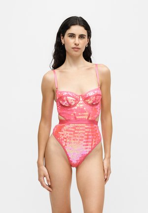 Femme aux longs cheveux foncés portant un maillot de bain une pièce rose brillant à sequins avec des découpes sur les côtés, debout devant un fond clair uni.