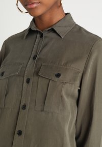 Close-up of person wearing olive green button-up shirt with two front flap pockets and gold hoop earrings.