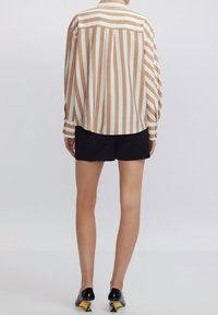 Camicia a maniche lunghe con righe verticali crema e beige, realizzata in tessuto leggero. Presenta una spalla a yoke e polsini con bottoni. Pantaloni corti neri e scarpe a punta.