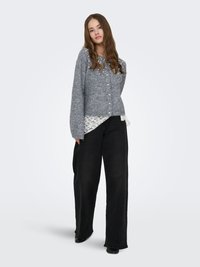 Cardigan grigio in maglia con dettagli in perle, chiusura con bottoni e design corto sovrapposto a una camicia bianca in pizzo, abbinato a pantaloni neri a gamba larga.