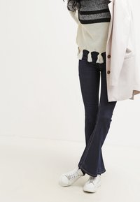 Pull en tricot rayé blanc et noir avec des franges, jean évasé bleu foncé, et baskets blanches. La personne tient un blazer rose clair.