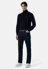 Maglione a collo alto blu navy, jeans blu scuro con cintura blu e sneaker bianche, esposti su uno sfondo bianco semplice.