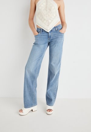 Kobieta ubrana w jasnoniebieskie szerokie jeansy, kremowy top na szydełku z wiązaniem na szyi oraz białe odkryte na palce obcasy, stojąca z rękami w kieszeniach.