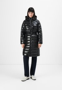 Donna che indossa un lungo cappotto imbottito nero lucido con cappuccio e cintura, jeans scuri e mocassini bianchi, in piedi contro uno sfondo bianco uniforme.