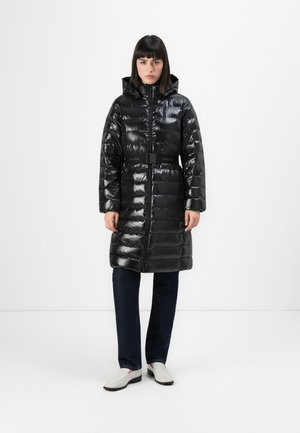Vrouw in een lange zwarte glanzende pufferjas met capuchon en riem, donkere spijkerbroek en witte loafers, staand tegen een effen witte achtergrond.