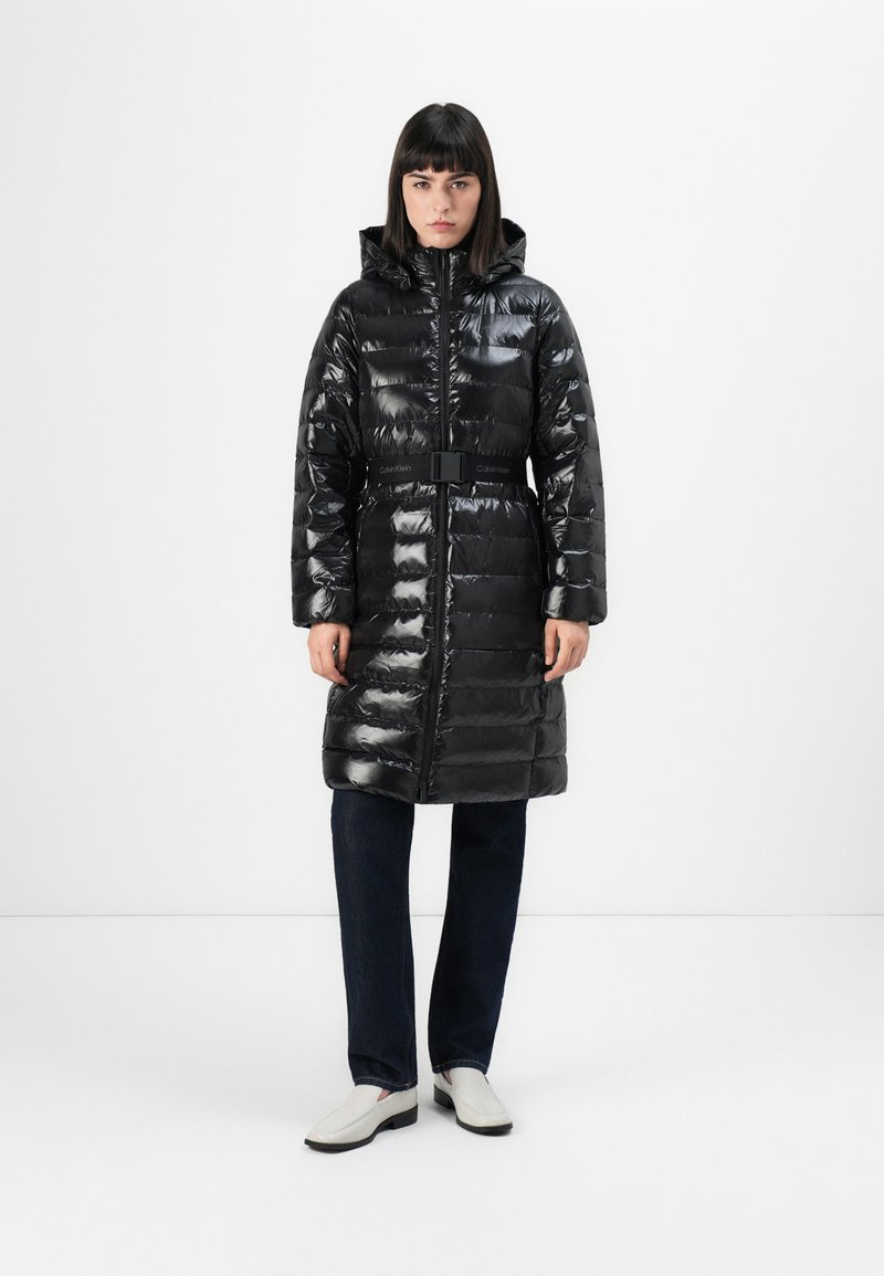 Donna che indossa un lungo cappotto imbottito nero lucido con cappuccio e cintura, jeans scuri e mocassini bianchi, in piedi contro uno sfondo bianco uniforme.