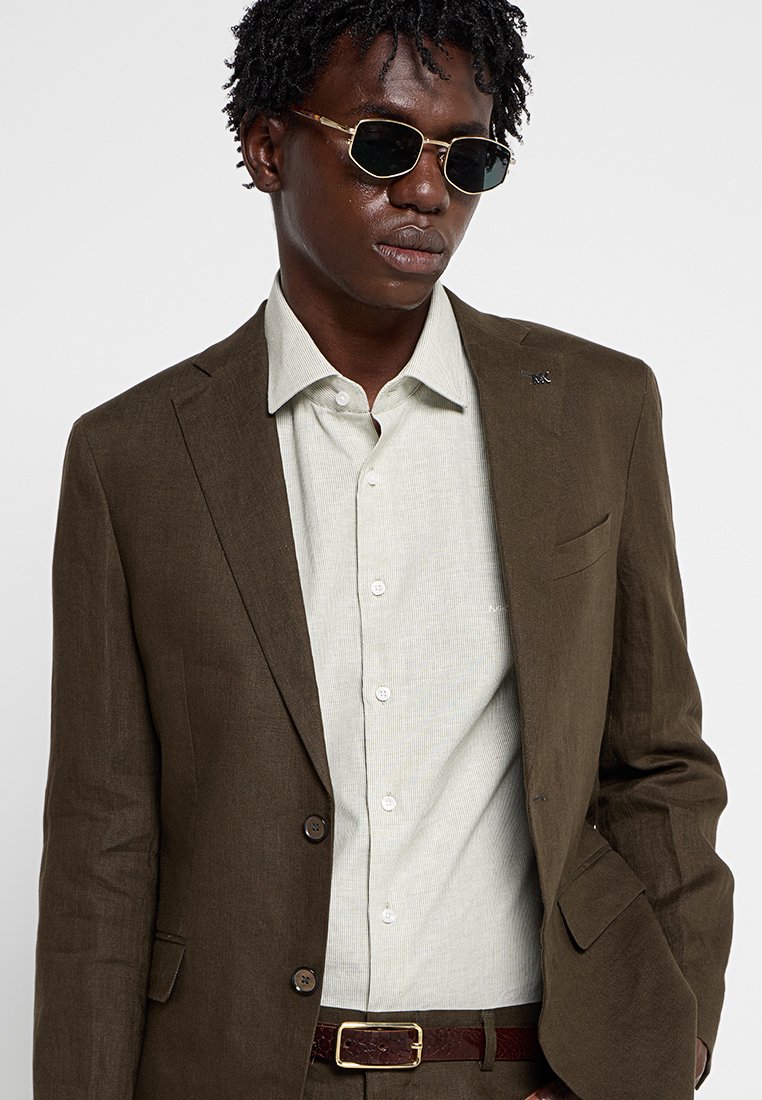 Brauner Leinenblazer über einem hellgrünen Hemd mit dunkler, gestreifter Textur. Das Outfit wird von einem dunkelbraunen Gürtel und dunklen Sonnenbrillen abgerundet.