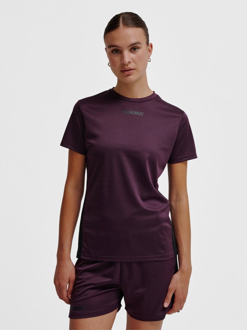Femme portant un t-shirt de sport manches courtes violet foncé de la marque hummel et un short assorti, debout sur un fond blanc.