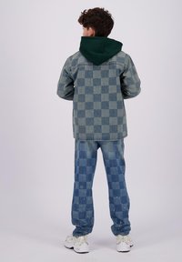 Giacca di jeans con cappuccio verde, caratterizzata da un motivo a quadretti. Pantaloni in denim a quadretti abbinati, di un blu chiaro con texture, abbinati a sneakers bianche.