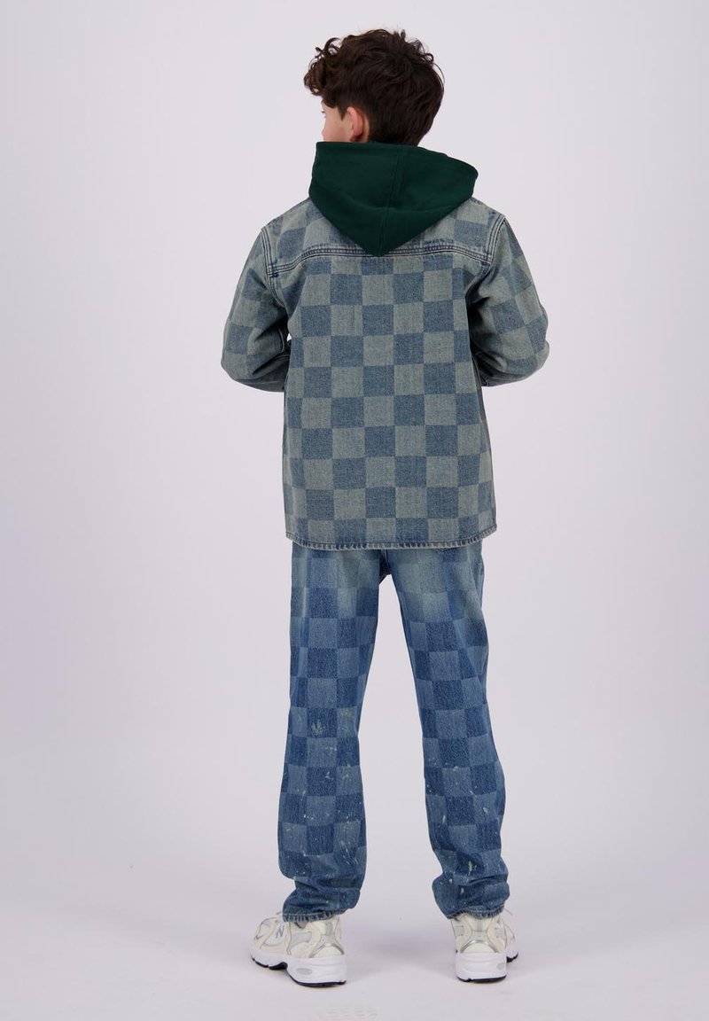Giacca di jeans con cappuccio verde, caratterizzata da un motivo a quadretti. Pantaloni in denim a quadretti abbinati, di un blu chiaro con texture, abbinati a sneakers bianche.