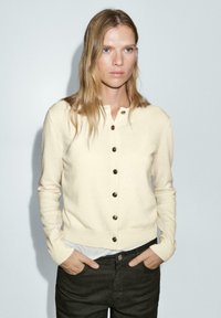 Cardigan crème en matière douce, avec un col rond, des boutons sur le devant et des poignets côtelés, associé à un pantalon sombre.