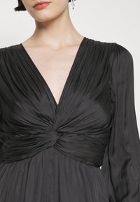 Femme portant une robe noire plissée avec un décolleté en V profond et un détail de nœud torsadé à la taille, associée à des boucles d'oreilles pendantes.