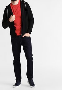 Sudadera negra con cremallera y cordones blancos, camiseta roja debajo, jeans azul oscuro y zapatillas azules. Un atuendo sencillo y casual.