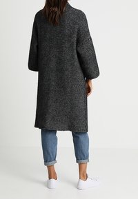 Lång svart och grå stickad cardigan med vida ärmar, texturerad tyg och öppen rygg, ihop med upprullade blå jeans och vita sneakers.