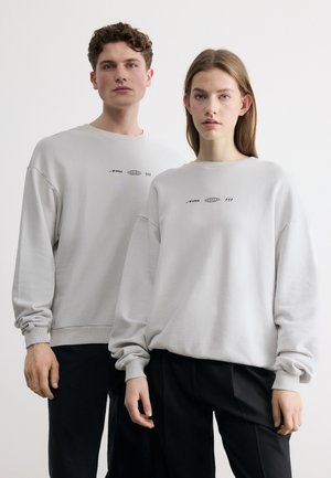 Młody mężczyzna i kobieta stojący obok siebie w pasujących jasnoszarych bluzach z małymi czarnymi logo, czarne spodnie, neutralne tło.