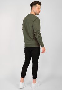 Olivgrön långärmad sweatshirt med ribbade manschetter, kombinerad med svarta slim-fit byxor och vita sneakers, sedd bakifrån.