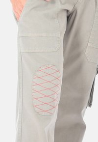 I pantaloni cargo grigi chiari presentano una toppa sul ginocchio con cuciture a zigzag rosse. Comprendono tasche laterali e un tessuto durevole e testurizzato.