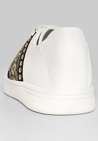 Zapatilla blanca con un talón liso, suela blanca y panel lateral de tela negra con tachuelas doradas y un bordado intrincado.
