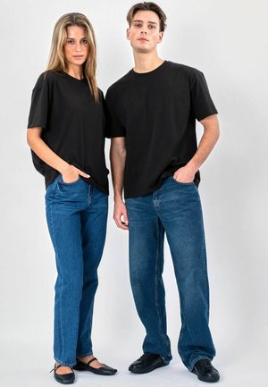 Kvinde og mand iført sorte t-shirts og blå jeans, stående side om side med hænderne i lommerne mod en ensfarvet baggrund.