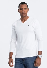 Weißes langärmeliges V-Ausschnitt-T-Shirt mit kleinem Logo auf der Brust, kombiniert mit blauen Jeans und einer silbernen Uhr am Handgelenk.