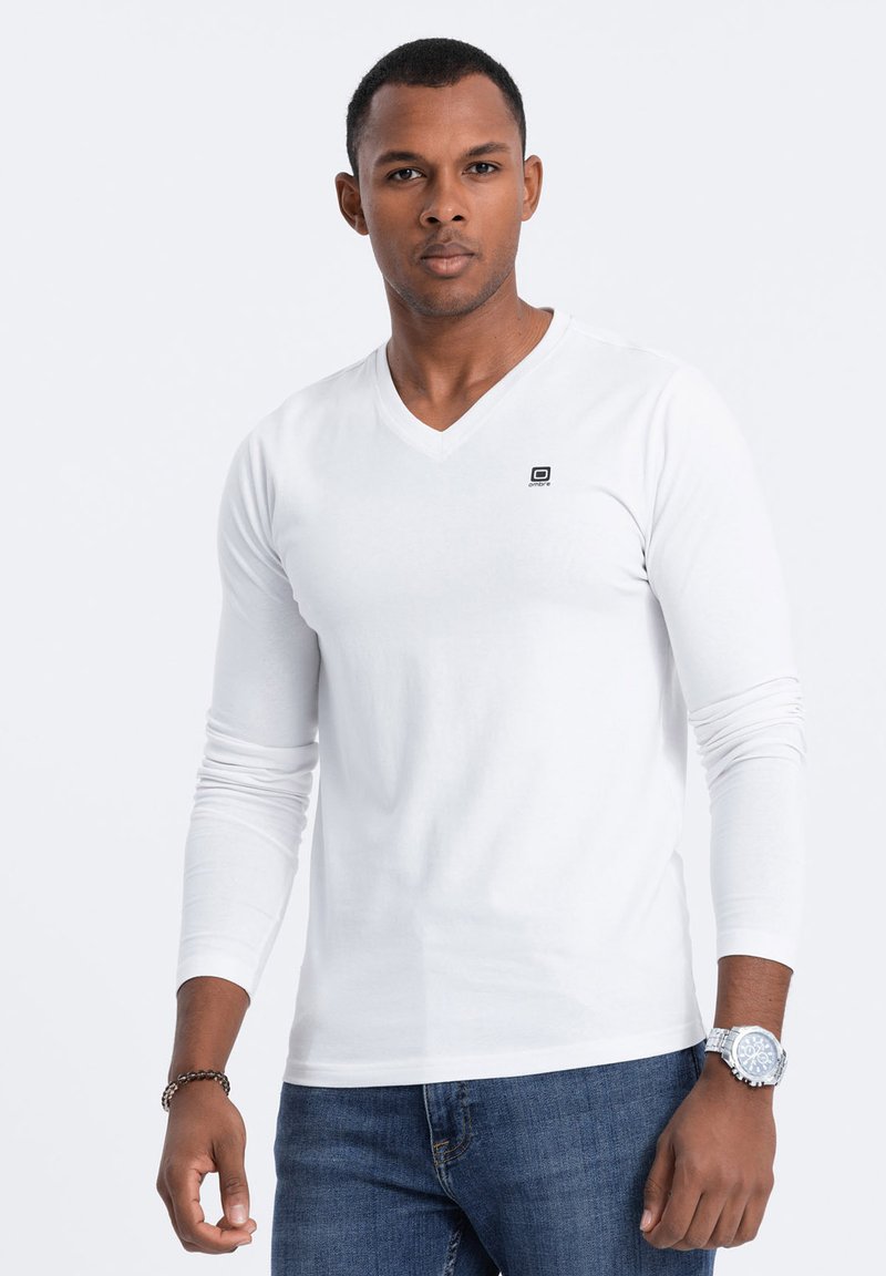 Weißes langärmeliges V-Ausschnitt-T-Shirt mit kleinem Logo auf der Brust, kombiniert mit blauen Jeans und einer silbernen Uhr am Handgelenk.