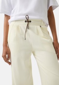 Pantalons crème avec une taille à cordon de serrage, dotés d'un cordon marron et de quincaillerie décorative. Design à plis sur le devant, forme à jambes larges.