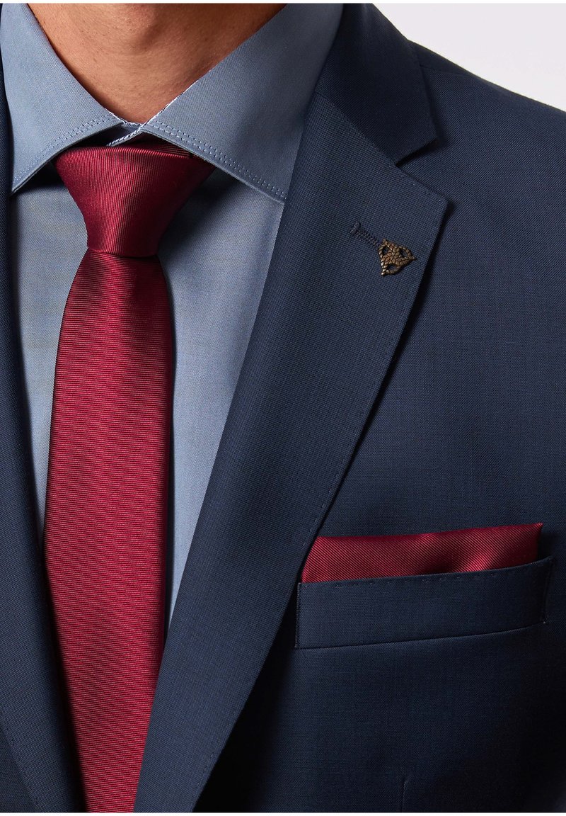 Giacca di abito blu navy con una camicia azzurro chiaro, cravatta rosso scuro e fazzoletto coordinato. Presenta un fermaglio decorativo sulla bavero. Texture liscia in tutto.