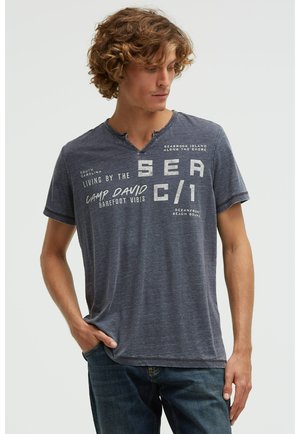 Young man with curly hair wearing a faded navy T-shirt with beach-themed text, standing with one hand in pocket, looking down left.