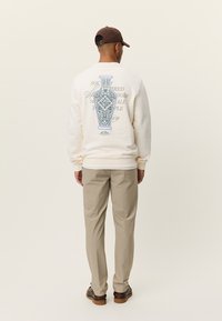 Μπλούζα φούτερ σε κρεμ απόχρωση με μπλε γραφικό βάζο στην πλάτη, μακριά μανίκια, ριμπ μανσέτες, συνδυασμένη με μπεζ παντελόνι και καφέ παπούτσια.