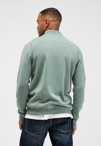 Groene gebreide trui met een hoge kraag, geribde manchetten en zoom. Gelaagd over een wit shirt, gecombineerd met donkerblauwe spijkerbroek.