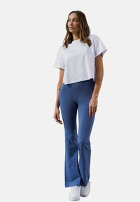 Biała krótka koszulka połączona z niebieskimi dżinsami w kształcie dzwonów; gładki materiał z minimalną teksturą; modelka w białych trampkach.