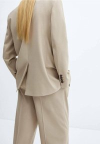 Beige Anzug, maßgeschneidert, mit einem einzelnen Schlitz, weiten Hosen und geknöpften Manschetten. Die weiche Textur verstärkt das anspruchsvolle Design.