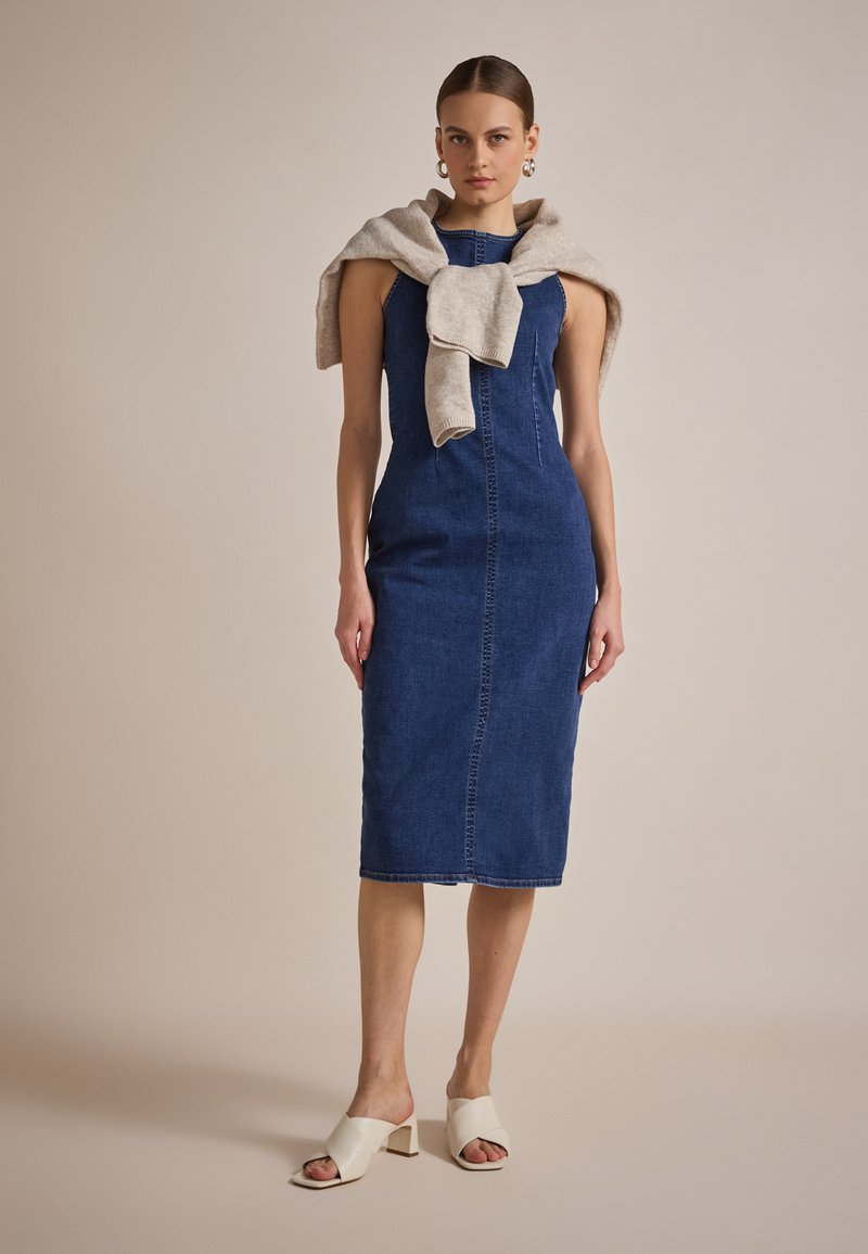 Mørkeblå denim midi-kjole med en tettsittende silhuett, som har en midtseam foran. En lys beige genser er drapert over skuldrene.