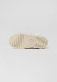 Såle av en sneakers som har en textured lys beige gummibunn med horisontale spor og et logo preget på hælområdet.