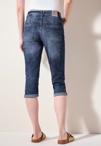 Pantalones capri de mezclilla en azul oscuro con una textura desgastada, puños enrollados y bolsillos traseros con detalles de costura. Etiqueta de cuero en la cintura.