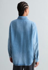 Person mit geflochtenem Haar, die eine hellblaue Jeansjacke trägt, von hinten vor einem schlichten hellgrauen Hintergrund gezeigt.