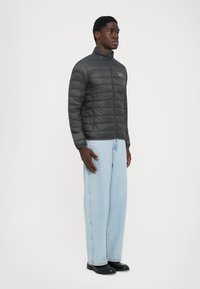 Mann står iført en svart quiltet jakke, lyseblå vide jeans og svarte dressko mot en ensfarget hvit bakgrunn.