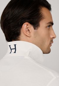 Biela polo košeľa s vysokým golierom, na zadnej strane s modrým logom "H". Hladká textúra a priliehavý strih.
