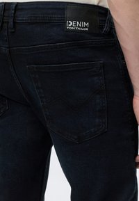 Detail zadného vrecka tmavomodrých džínsov s čiernou štítkom s nápisom "DENIM TOM TAILOR" na páse, nosených osobou v bielej košeli.