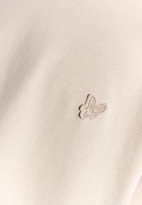 Beige tekstil med en tekstureret overflade og et subtilt, broderet sommerfugle-logo på venstre side.