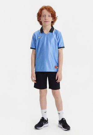 Garçon aux cheveux roux portant un maillot de sport Nike bleu clair, un short noir, des chaussettes Nike blanches et des baskets Nike noires sur fond blanc.