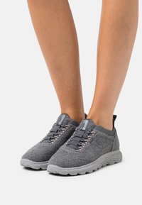 Grijze textielsneakers met een ronde neus, elastische veters en een lichte grijze rubberen zool. Kenmerken zijn een gebreide kraag en branding op de tong.